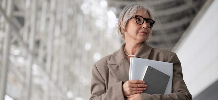 The middle-aged businesswoman, dressed in a professional outfit, thoughtfully gazes ahead while holding a laptop and notebook in a contemporary office environment.