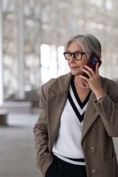 An elderly woman with a trendy look speaks on her phone while standing in a bright, contemporary space. She exudes confidence and style with her fashionable attire.