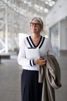 A senior businesswoman exudes confidence while standing outdoors. She holds a tablet in one hand and a coat in the other, dressed elegantly, ready for her next meeting.