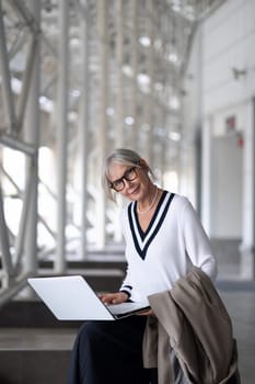 An elderly businesswoman sits at a desk, focused on her laptop in a contemporary office setting filled with natural light. She wears a stylish outfit and glasses, exuding professionalism.