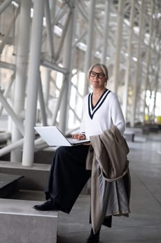 A fashionable elderly woman with blonde hair sits casually on a ledge, engaged in work on her laptop while holding a coat, surrounded by sleek architecture and natural light.