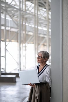 A middle-aged businesswoman stands against a wall in a contemporary office, holding a laptop while contemplating her next steps. Natural light fills the space.