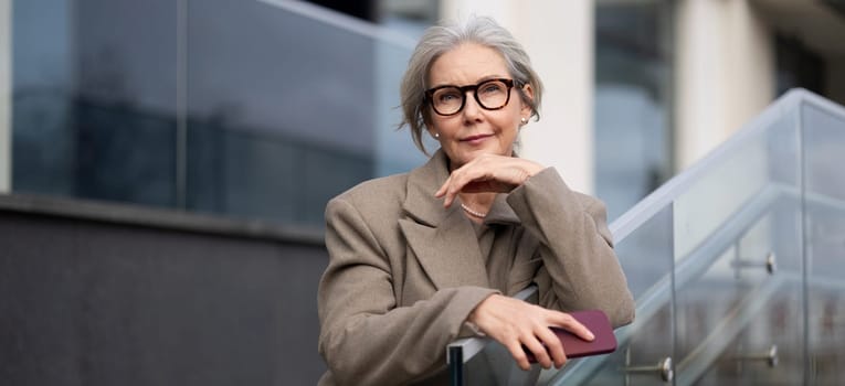An older woman with gray hair is leaning on a railing. She wears stylish glasses and a beige coat, thoughtfully gazing into the distance outside a contemporary building.