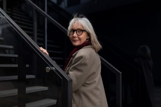 An elderly businesswoman with glasses and gray hair stands on the stairs outside a contemporary office building, exuding confidence and poise.