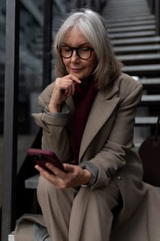A middle-aged businesswoman is seated on steps in an urban environment, intently using her smartphone. She appears thoughtful, dressed in a stylish coat and glasses.
