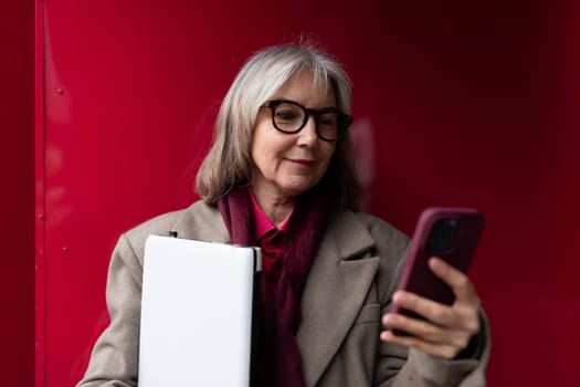 An elderly businesswoman stands outside her office, dressed in professional attire. She is intently looking at her phone while holding a notebook, showcasing her commitment to work.