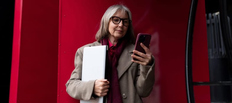 A professional senior businesswoman stands outdoors, examining her smartphone intently while clutching a portfolio. The bright red backdrop adds a modern touch to her composed demeanor.