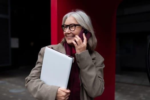 A middle-aged Caucasian businesswoman enjoys a cheerful phone conversation while holding a folder. She stands outside an office building, radiating confidence and approachability.
