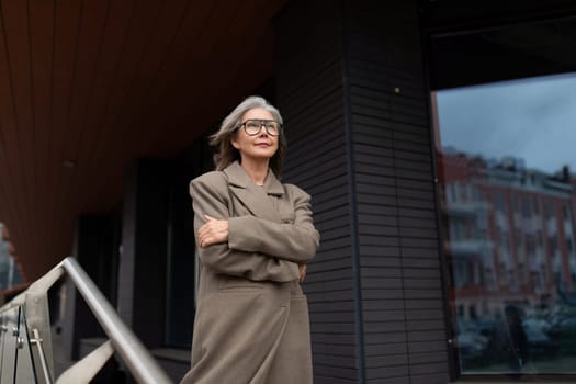 An older woman wearing a fashionable coat stands outside a contemporary building, arms crossed and exuding confidence. She enjoys a sunny day, looking at the surroundings.