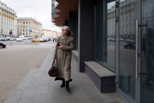 A confident elderly businesswoman strolls along a busy city street, wearing a stylish coat and carrying a handbag, displaying poise and professionalism.