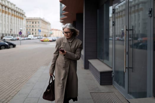 A senior businesswoman walks along the sidewalk, engrossed in her phone. She wears a stylish long coat and carries a handbag, showcasing an urban lifestyle.
