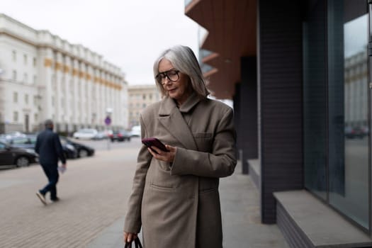 An elderly businesswoman dressed in a stylish coat stands on the sidewalk, focused on her smartphone, as she walks past an office building on a gloomy day.