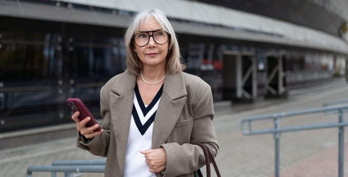 An elderly businesswoman in a stylish outfit holds her phone as she stands outside a modern office building, enjoying the sunny day while looking thoughtful.