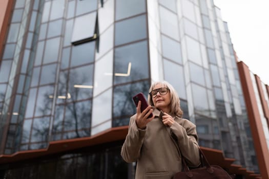 A senior businesswoman stands outside a contemporary office building, focused on her smartphone. She wears a stylish coat and holds a bag, appearing engaged in her task.