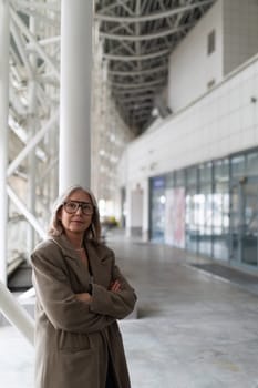 An elderly businesswoman poses confidently in a spacious, modern urban area, arms crossed, reflecting her experience and professional outlook in a contemporary setting.