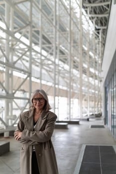 An elderly businesswoman exudes confidence as she stands in a bright, contemporary office space filled with natural light. Her presence highlights her experience.