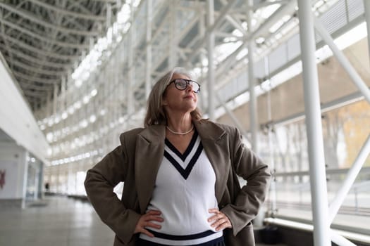 The older woman stands tall with her hands on her hips, exuding confidence in a spacious, well-lit area filled with modern architecture. Her attire reflects a sophisticated style.