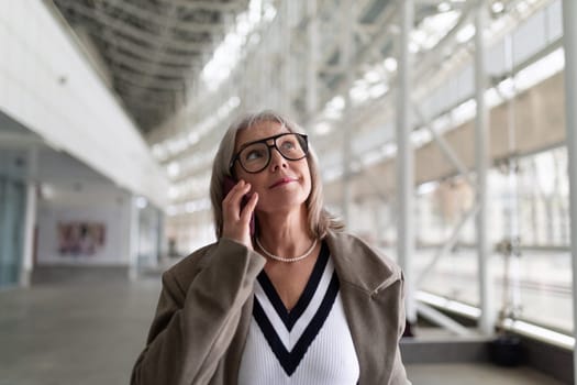 An older woman stands in a contemporary indoor space, her thoughtful expression highlighting her style. She looks up as if contemplating the surroundings, showcasing her confidence.