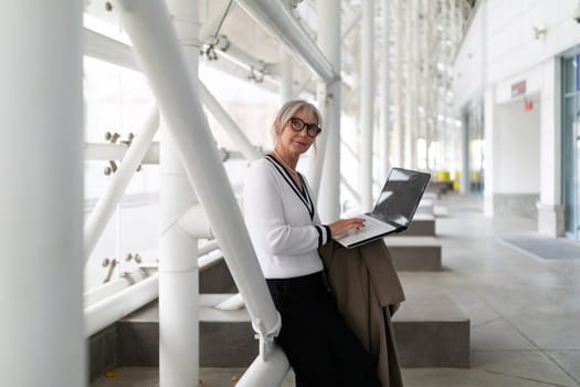An elderly businesswoman is focused on her laptop, sitting comfortably in a modern office environment.