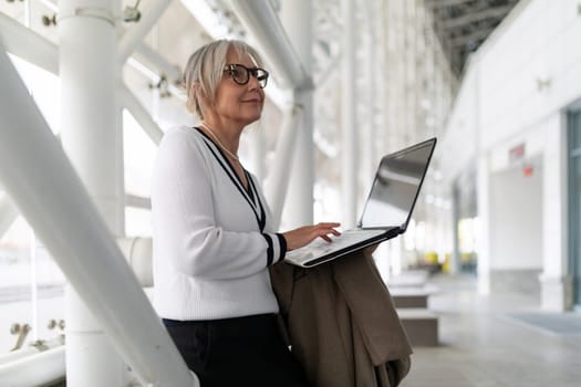 A fashionable elderly woman stands by a pillar, focused on her laptop while holding a coat, in a contemporary indoor environment filled with light.