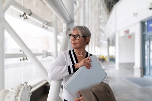 An elderly businesswoman stands outside her office, holding a laptop bag close to her chest. She gazes thoughtfully, dressed in professional attire, exuding confidence.