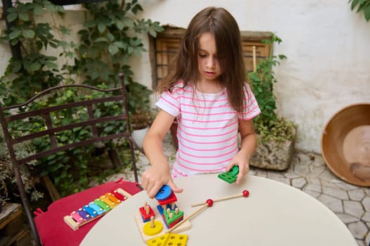 A young girl plays with colorful educational toys on a table in a garden. Her focused expression and the serene outdoor atmosphere create a peaceful and educational environment.
