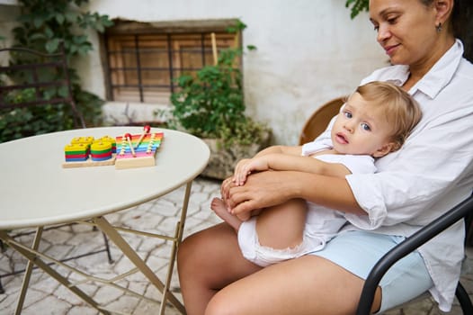 A mother holds her young child on her lap while sitting outdoors. Colorful toys are placed on a nearby table, creating a serene and family-oriented atmosphere at home.