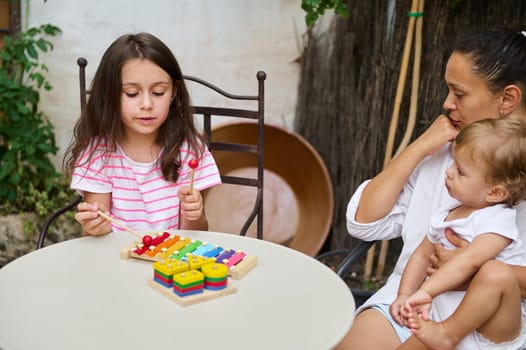 A young girl plays a colorful xylophone on a table outside, as her mother attentively watches while holding a baby. The family shares a joyful moment in a cozy outdoor setting.