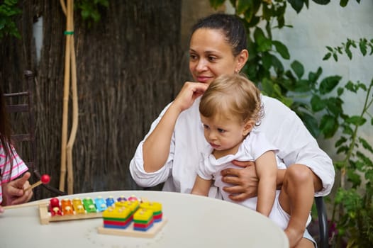 A mother lovingly holds her child while watching colorful toys on a table outside. The scene exudes warmth, togetherness, and maternal bonding in a natural garden setting.