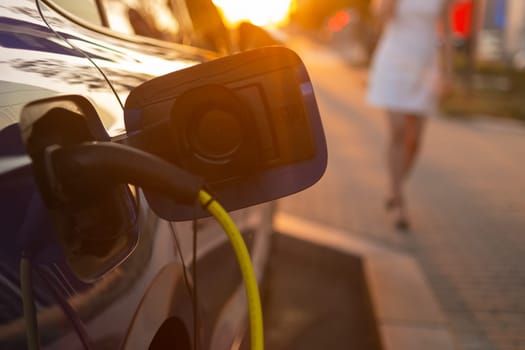 Close up electric car charging, while a woman in a white dress is walking beside it at sunset.