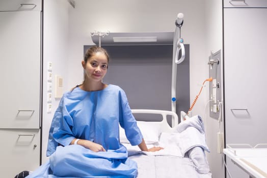 Young female patient in hospital ward,dressed in blue gown,sitting on bed and waiting to be examined by doctor,smiling softly and satisfied with service and services provided,individual hospital room