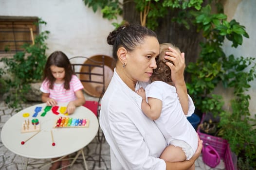 A thoughtful mother holds her baby while her older child plays with colorful toys in a garden setting, embodying family love and quiet reflection.