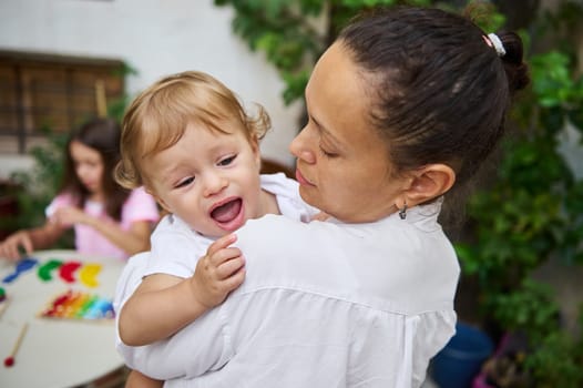 A loving mother holds her joyful baby while another child plays with colorful toys on a table in the garden.