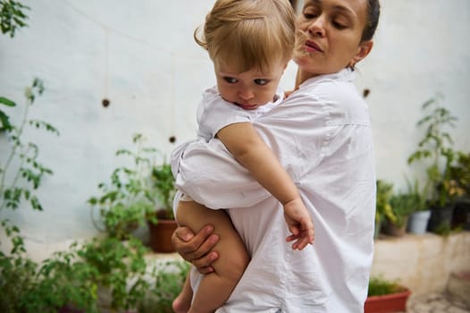 A mother lovingly cradles her young child in an outdoor garden, surrounded by lush greenery, portraying a tender moment of familial bond and care.