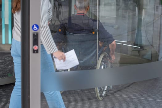 middle-aged man sitting in a wheelchair moves towards the glass revolving door of a clinic, using his hands to control the wheels, his wife follows behind, holding the patient's medical record,
