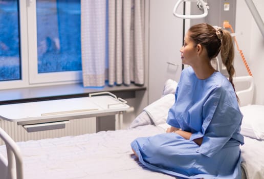 young woman in a private hospital room, dressed in a blue gown, sits on a bed and waits to be examined by a doctor before plastic surgery,