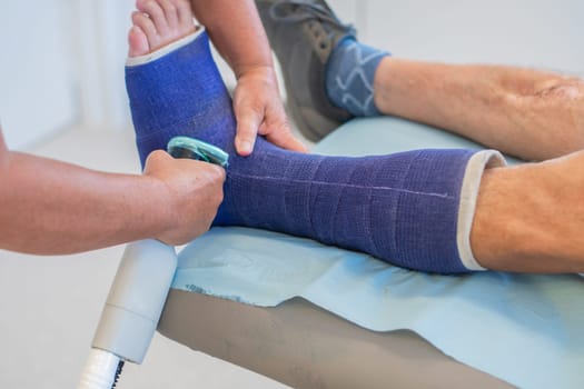 Nurse in clinic carefully cuts strong fiberglass cast on patient's leg with plaster saw according to instructions, taking precautions, patient sits quietly, professional help concept,