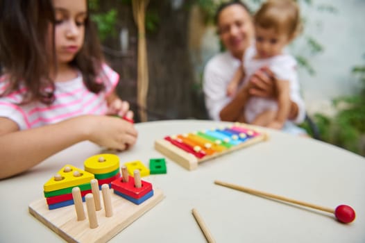 A young child engaging with colorful educational wooden toys at a table, while a family scene unfolds in soft focus. Emphasis is on learning and play.