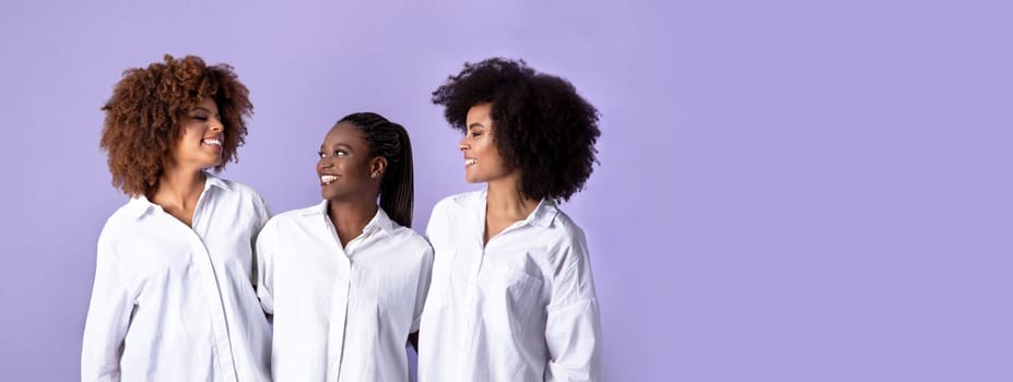 Beauty concept. Happy three african american ladies embracing and smiling to each other, posing wearing shirts over purple background, studio shot of sisters or friends, women trio