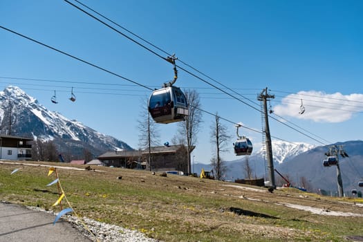 Cable cars glide above mountainous terrain on a clear day, offering views of snowy peaks and green pathways near a ski resort - Rosa Khutor, Russia April 15, 2023.