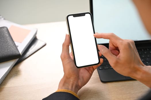 Businessman holding smartphone with empty screen sitting in front of digital tablet at desk.