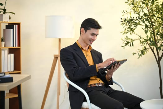 Asian businessman in black suit analyzing data on digital tablet in modern workplace.