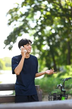 Smiling asian man talking on mobile phone while standing near his bicycle in the park.