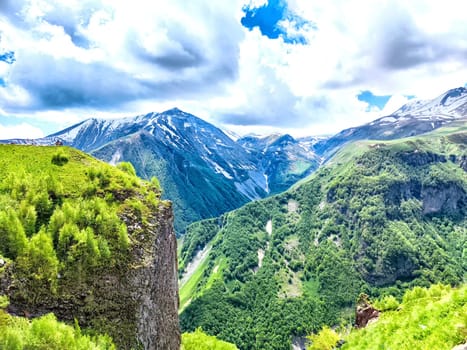 A vast view of mountains featuring greenery and snow under a bright, partly cloudy sky.