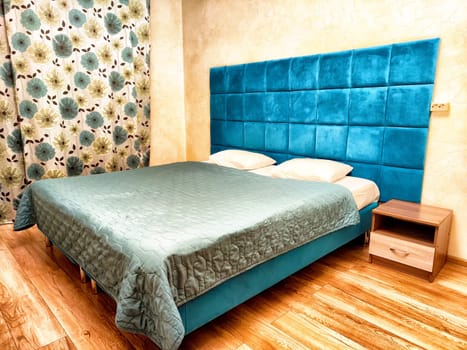 The cozy bedroom features a teal upholstered headboard, soft bedding, and a floral curtain, creating a calming atmosphere.