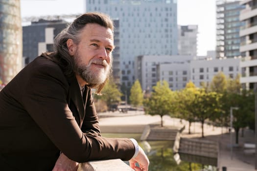 positive middle age male with gray hair and beard in stylish formal outfit looking away pensive while leaning on fence on terrace of city building, concept of business and mature man lifestyle