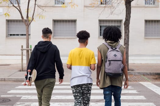 rear view of three diverse teen friends with backpack and skateboard crossing road on crosswalk, concept of youth and urban lifestyle