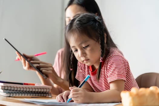 A young girl focused on drawing with colored pencils as her mother works on a tablet, highlighting creativity and family time at home.