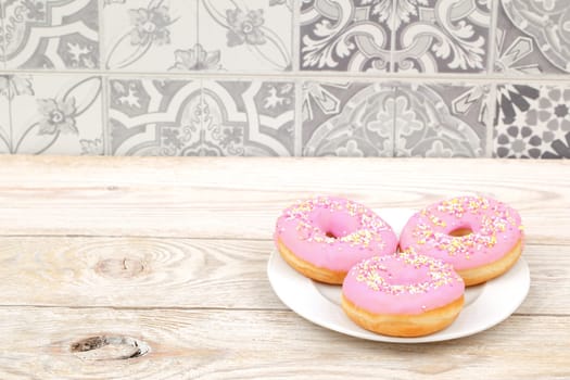 Three pink frosted donuts with colorful sprinkles, arranged on a white plate against a stylish patterned background