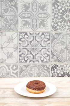 A tempting chocolate donut sits on a white plate, surrounded by a beautifully decorative tile background
